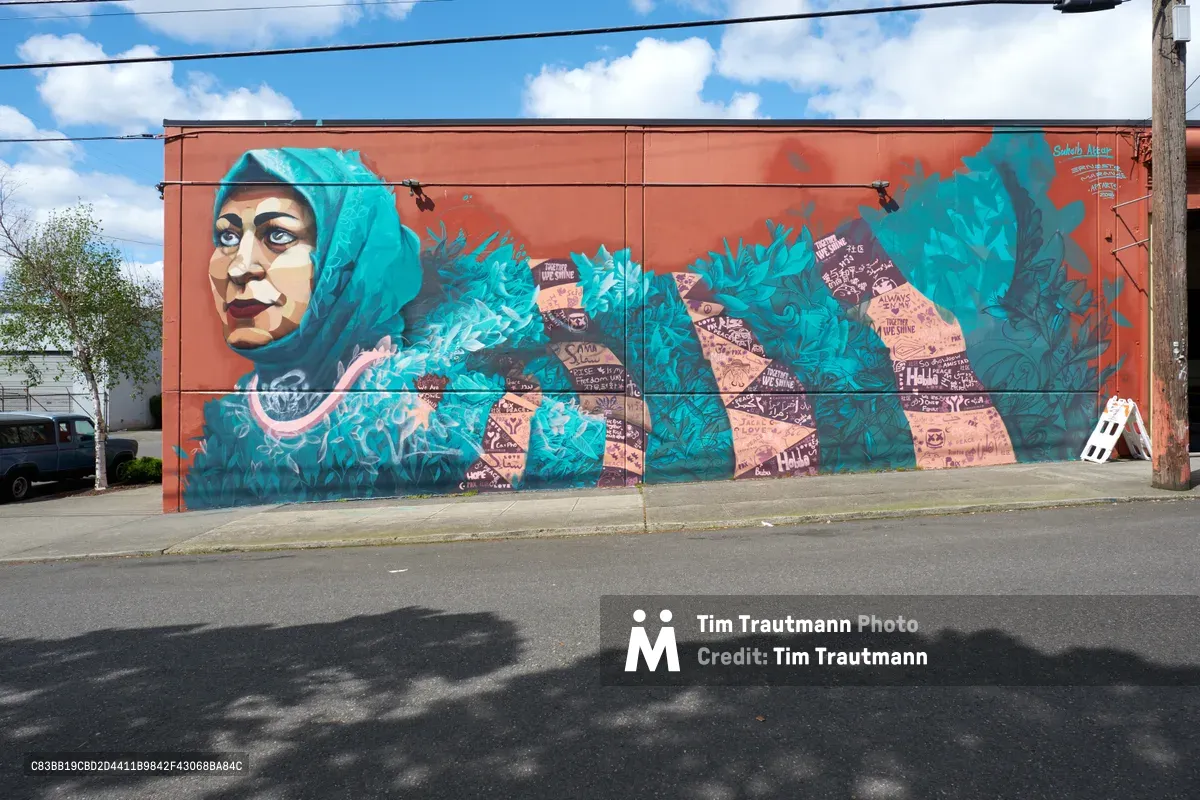A commanding street mural dominates a rust-colored building wall in Portland's Central Eastside district, featuring a woman's portrait rendered in luminous turquoise and coral tones. The artwork, created by Suhaib Attar and Ernesto Maranje for aptART, flows across the brick facade with swirling organic forms and embedded text fragments that speak to community resilience. Power lines cut across the azure sky dotted with cotton-white clouds, while concrete sidewalk and asphalt street frame the urban canvas below.