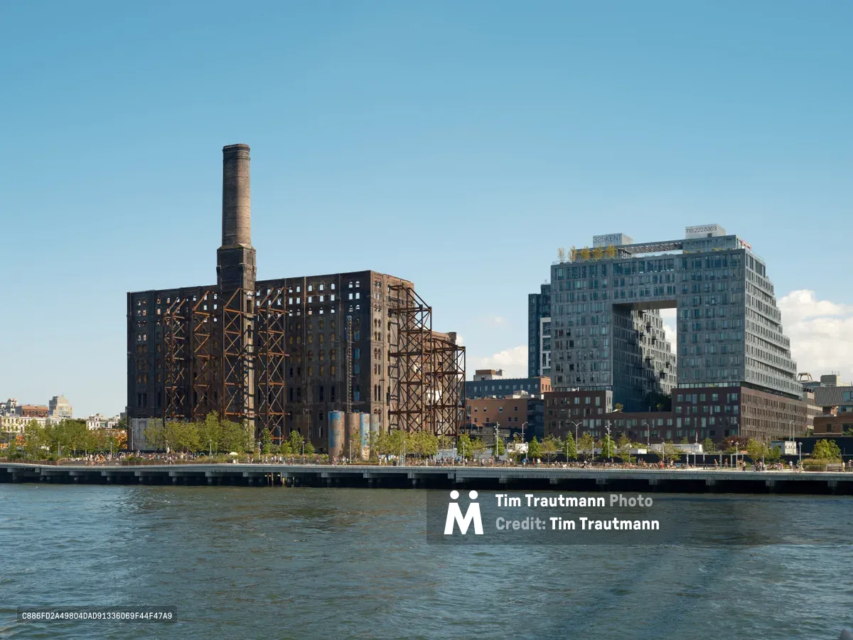The skeletal remains of the historic Domino Sugar Factory stand sentinel against a cloudless blue sky, its weathered brick facade and exposed steel framework creating a dramatic contrast with the sleek glass towers of 325 Kent Avenue rising behind it. The East River flows in the foreground, reflecting the afternoon light as pedestrians stroll along the Williamsburg waterfront promenade. This striking juxtaposition captures Brooklyn's ongoing transformation from industrial past to contemporary urban landscape.