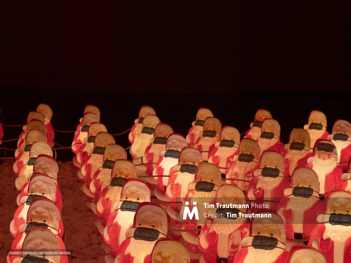 Dozens of identical Santa Claus figures wearing black face masks stand in neat military-like rows against a deep burgundy backdrop in Portland's Westmoreland neighborhood. The installation by artist Chris Willis transforms the jolly holiday icon into a haunting commentary on pandemic-era isolation, with warm amber lighting casting shadows across the mass-produced faces. Each figure maintains the classic red suit and white beard while the stark black masks create an unsettling uniformity that speaks to collective experience during COVID-19.