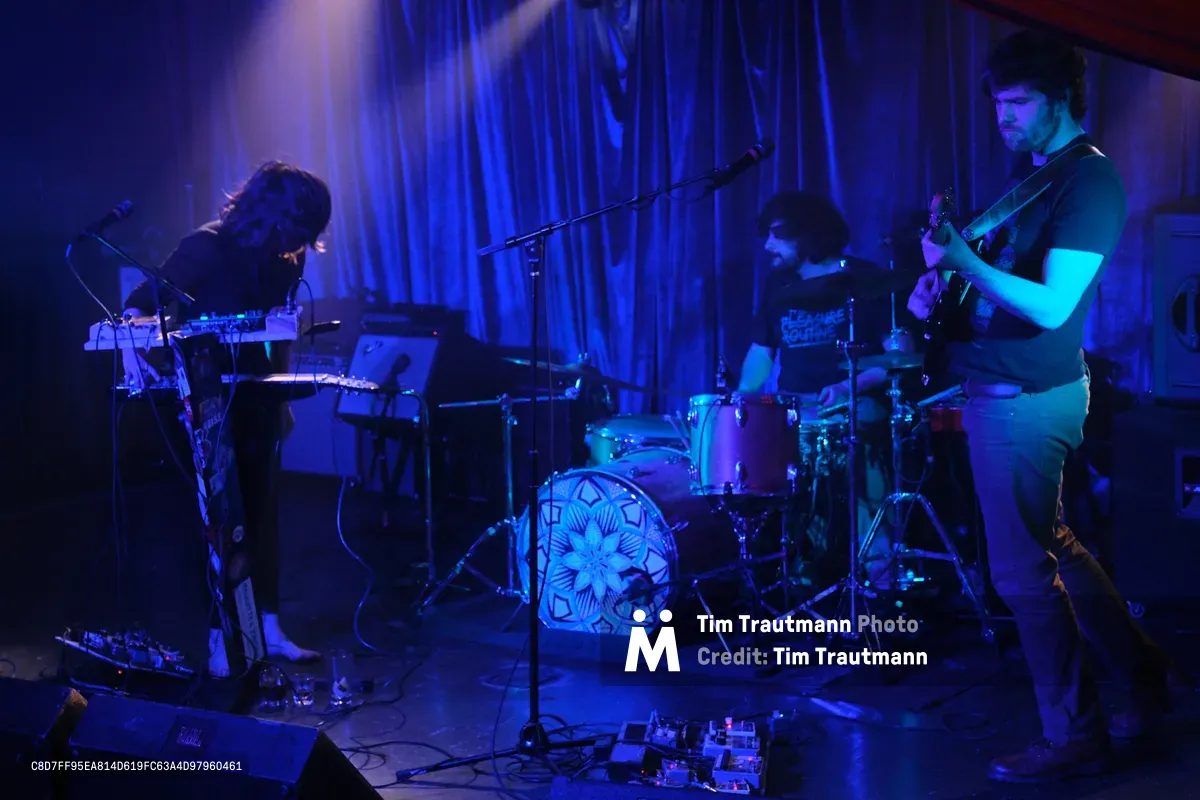 Boise experimental band Sun Blood Stories delivers an atmospheric performance at Rx Fest within the intimate confines of Dante's in downtown Portland. Deep purple and blue stage lighting bathes the three-piece band as they navigate their sonic landscape - the keyboardist hunched over synthesizers on the left, drummer maintaining rhythm behind a geometric kick drum design, and bassist anchored on the right. The moody illumination transforms the small venue into a nocturnal sanctuary where electronic textures merge with live instrumentation, creating the mesmerizing sound that has been likened to modern-day opera.