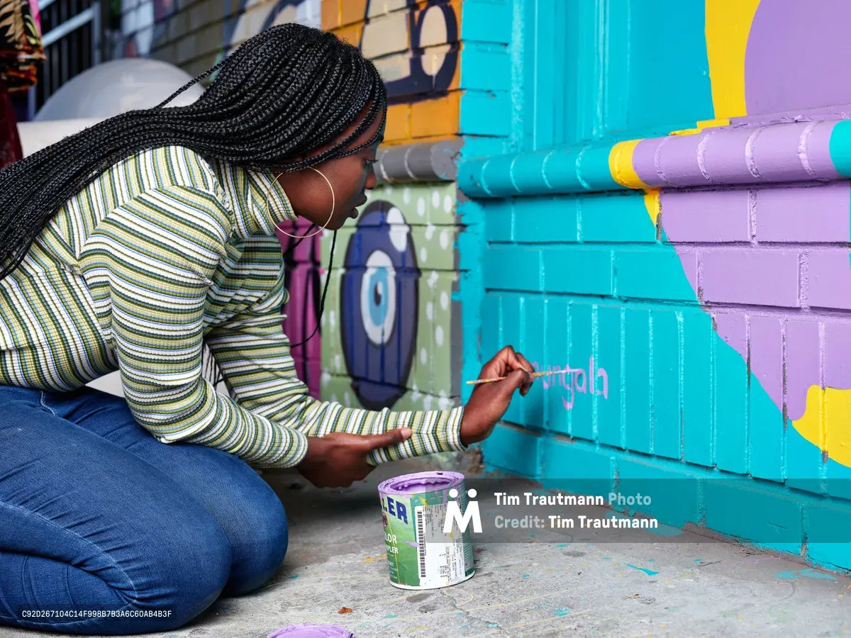 Naomi Likayi kneels on the sidewalk in Alberta Arts District, carefully painting her signature in purple letters across the vibrant turquoise and lavender geometric mural behind her. Her intricate box braids frame her concentrated expression as she works with a small brush, a paint can beside her on the concrete. The warm afternoon light illuminates her striped long-sleeve shirt and the bold street art that transforms this Northeast Portland corner into a canvas of community expression.