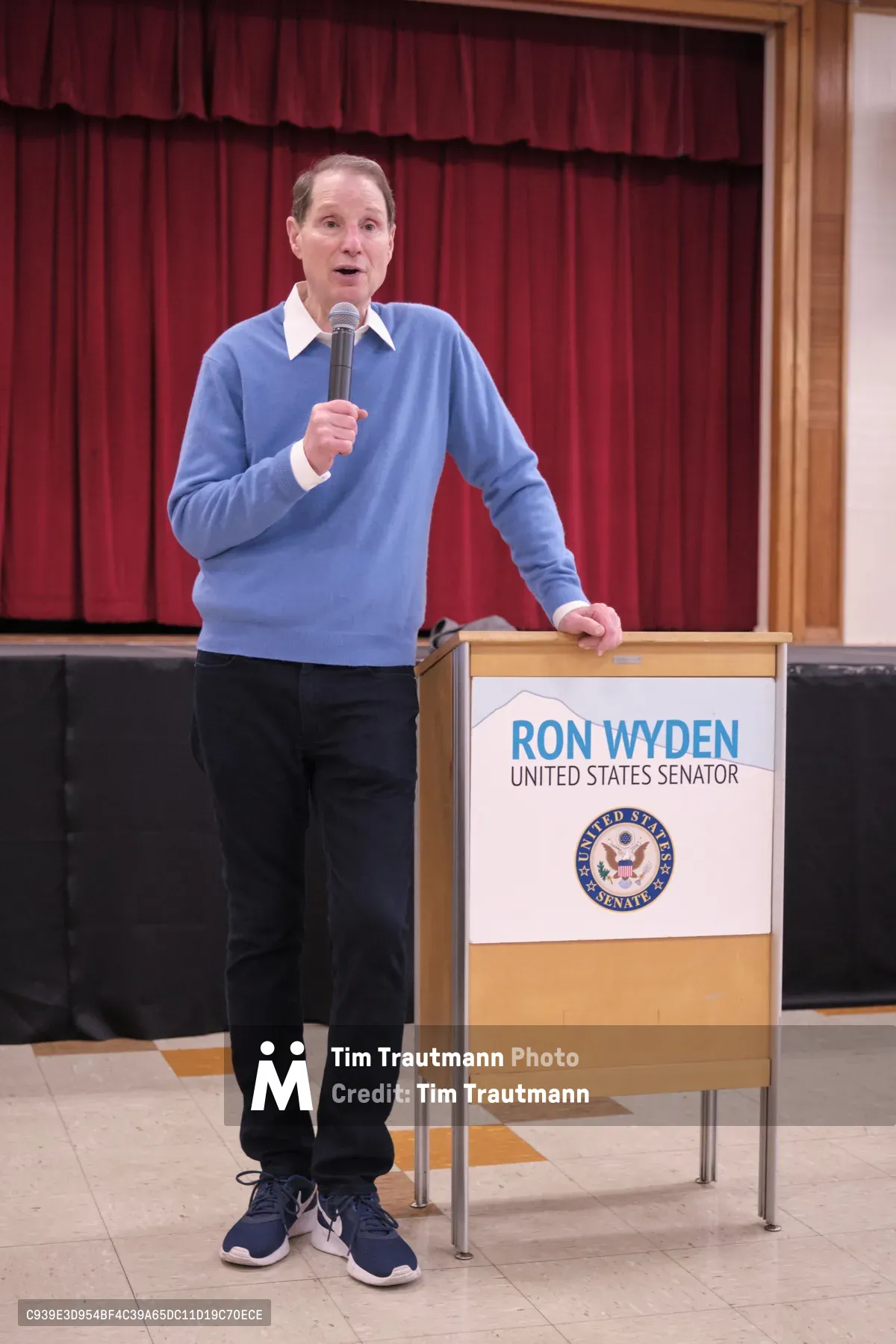 U.S. Senator Ron Wyden stands at a podium in the gymnasium of Robert Gray Middle School in Portland's Hillsdale neighborhood, addressing constituents during a town hall meeting. The senator, dressed casually in a blue sweater over a white collared shirt, grips a microphone while speaking against the backdrop of deep burgundy curtains that frame the school's performance space. The warm wooden podium bearing his official senatorial seal creates an intimate setting for this grassroots political engagement.