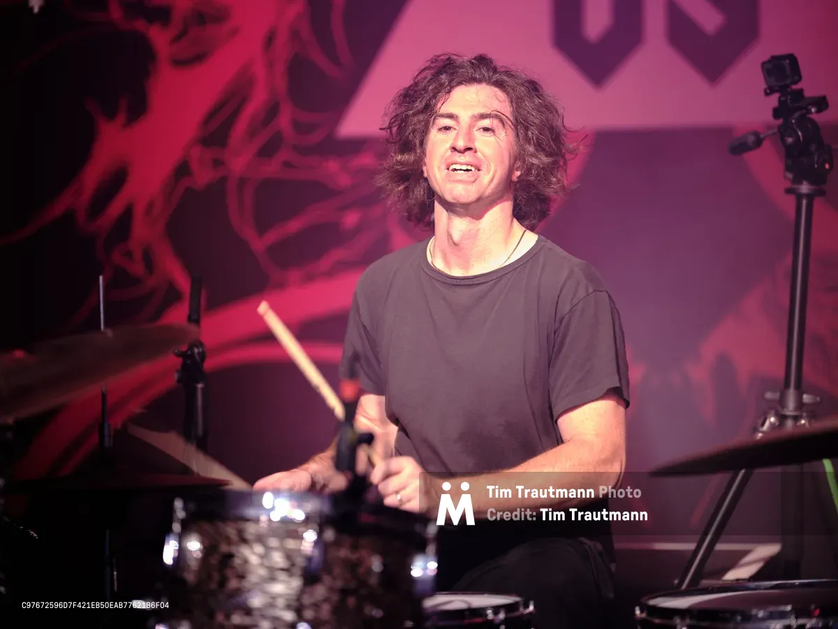 Julian Dorio, drummer for Band of Skulls, delivers an electrifying performance behind his kit at Manhattan's iconic Webster Hall during the band's 10 Year Anniversary Tour. Bathed in crimson stage lighting that transforms the intimate venue into a pulsing concert cathedral, Dorio's wild curls frame his concentrated expression as he grips his drumsticks mid-performance. The atmospheric lighting creates dramatic shadows across his simple black t-shirt while chrome cymbal surfaces catch and scatter the theatrical illumination throughout the storied East Village venue.