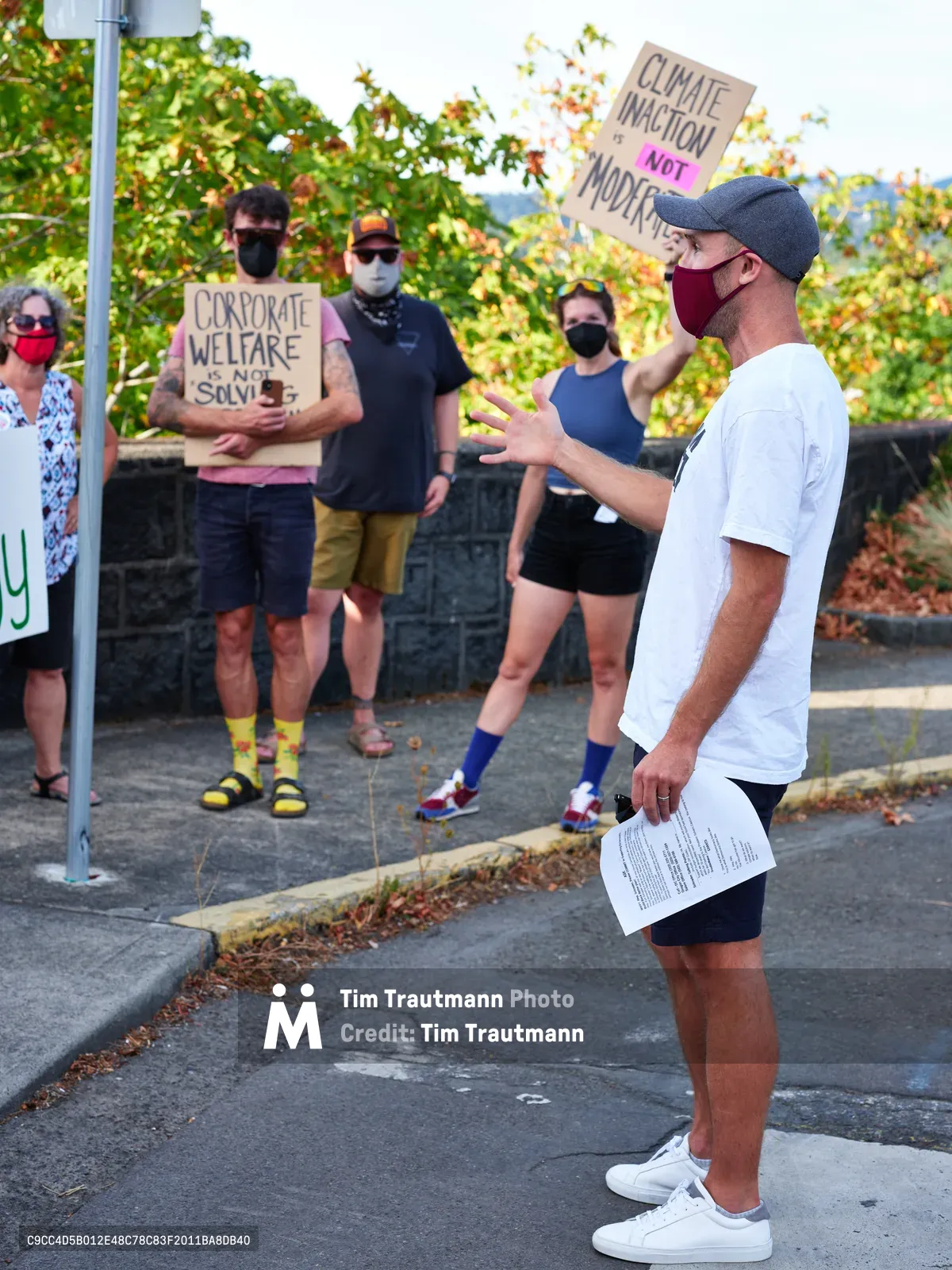 Peter Marks, wearing a red mask and baseball cap, gestures emphatically while addressing a small gathering of masked protesters outside Representative Kurt Schrader's Oregon City office. Behind him, activists hold handmade cardboard signs reading "CLIMATE INACTION NOT MODERATE" and "CORPORATE WELFARE IS NOT SOLVING," their colorful socks and casual attire contrasting with the serious political messaging. Golden autumn light filters through leafy trees, casting warm tones across the concrete sidewalk where this intimate act of civic engagement unfolds.