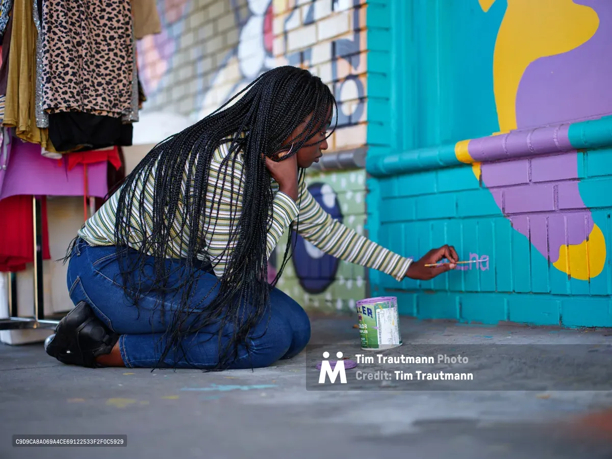 Naomi Likayi kneels on weathered pavement to sign her completed mural, her long braids cascading forward as she delicately applies her signature to the turquoise and purple brick wall. The scene captures an intimate moment of artistic completion in Portland's Alberta Arts District, where colorful murals transform urban spaces. Afternoon light illuminates the vibrant street art backdrop, creating a celebration of community creativity and cultural expression.