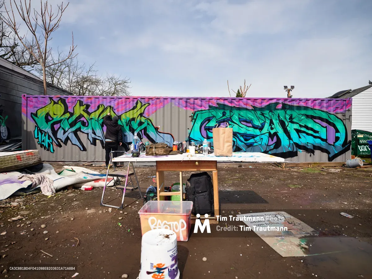 A makeshift artist's studio sits before a sprawling graffiti mural on a corrugated metal wall in Northeast Portland, Oregon. The vivid artwork features turquoise and purple wildstyle lettering that flows across the industrial surface, while paint supplies, canvases, and a wooden easel occupy the foreground under overcast winter skies. Bare branches and residential buildings frame this urban creative space, capturing the raw authenticity of street art culture in the Pacific Northwest.