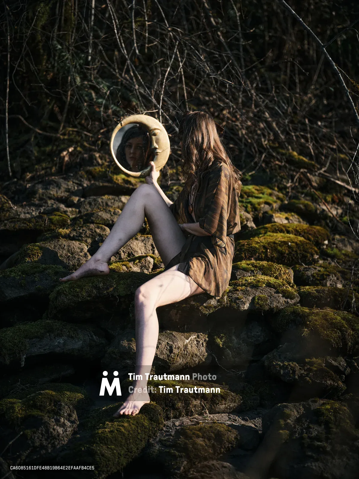 A contemplative figure sits barefoot on moss-covered rocks along a riverbed section of the Floodplain Trail in Multnomah County, Oregon, holding a circular mirror that captures their reflection. The subject wears an earth-toned jacket and dark leggings, positioned among weathered stones carpeted with vibrant green moss. Behind them, a dense tangle of bare winter branches creates a natural backdrop, while dappled sunlight filters through the forest canopy, casting warm highlights across the serene woodland scene.