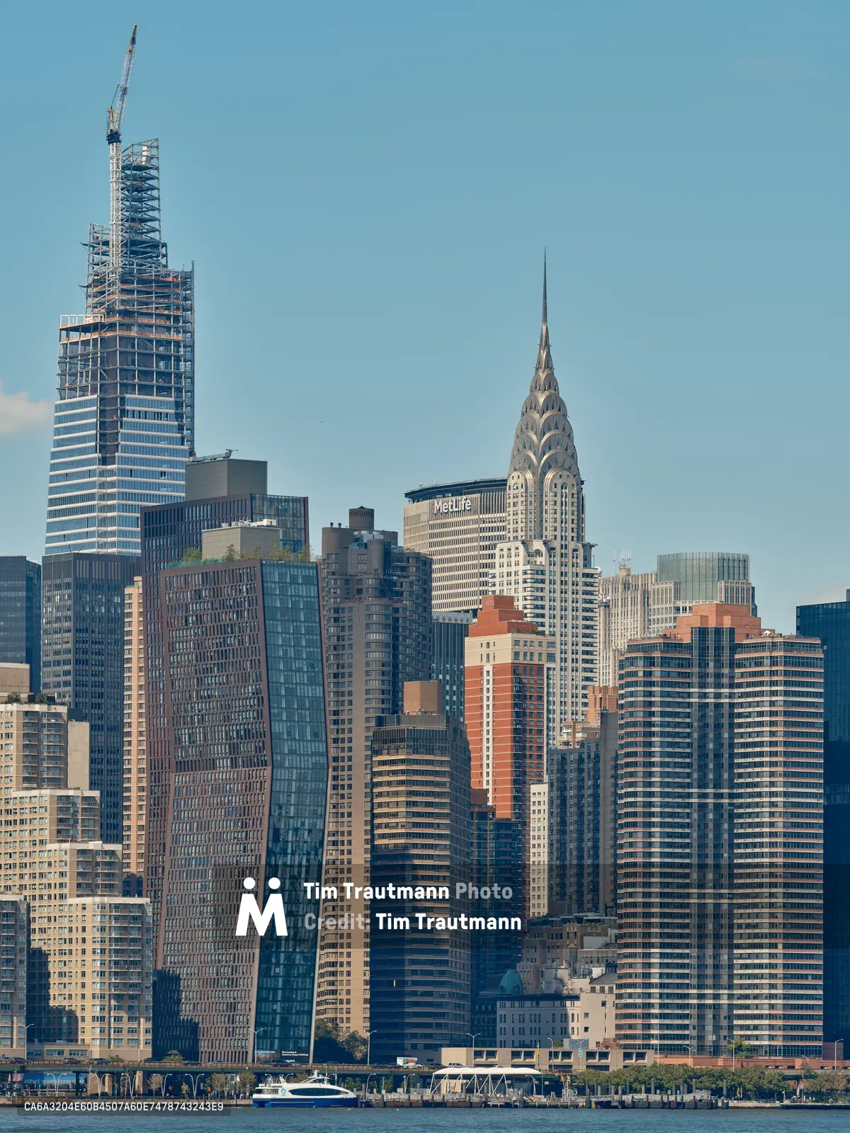 The Midtown Manhattan skyline viewed from across the East River under a clear blue sky. One Vanderbilt tower is visible under construction on the upper left, its steel frame topped with a construction crane. The iconic Art Deco Chrysler Building with its distinctive stainless steel eagle crown and spire rises prominently in the center of the skyline. The MetLife Building is visible behind it. A cluster of modern glass and brick residential and commercial towers fills the dense skyline. A ferry and waterfront promenade are visible along the river's edge in the foreground.