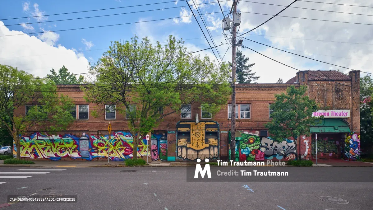 Vibrant street murals sprawl across the brick facade of the former Oregon Theater building on Southeast Division Street in Portland's Richmond neighborhood. The collaborative artwork, featuring bold geometric patterns, character portraits, and stylized lettering, creates a kaleidoscope of color against the weathered red brick under a dramatic cloudy sky. Power lines crisscross overhead while mature trees frame the scene, capturing the intersection of urban art and neighborhood history in this gentrifying corridor.