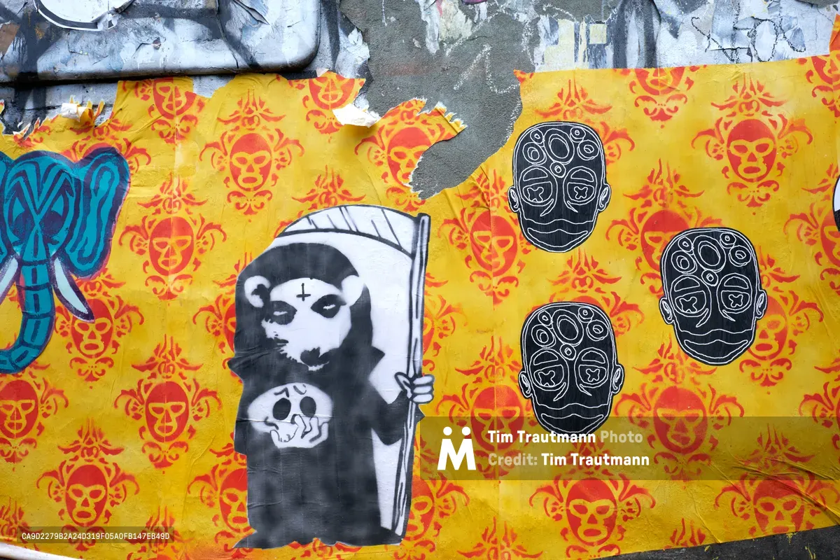 Weathered wheat paste murals blanket a concrete wall in Portland's Central Eastside, where mortality meets political commentary. A hooded Grim Reaper figure wielding a scythe dominates the left panel, juxtaposed against ornate sugar skull motifs scattered across vibrant yellow and red backgrounds. Three pristine white skulls with geometric patterns float like spectral guardians, their clean lines contrasting sharply with the deteriorating urban canvas beneath layers of peeling paint and exposed concrete.