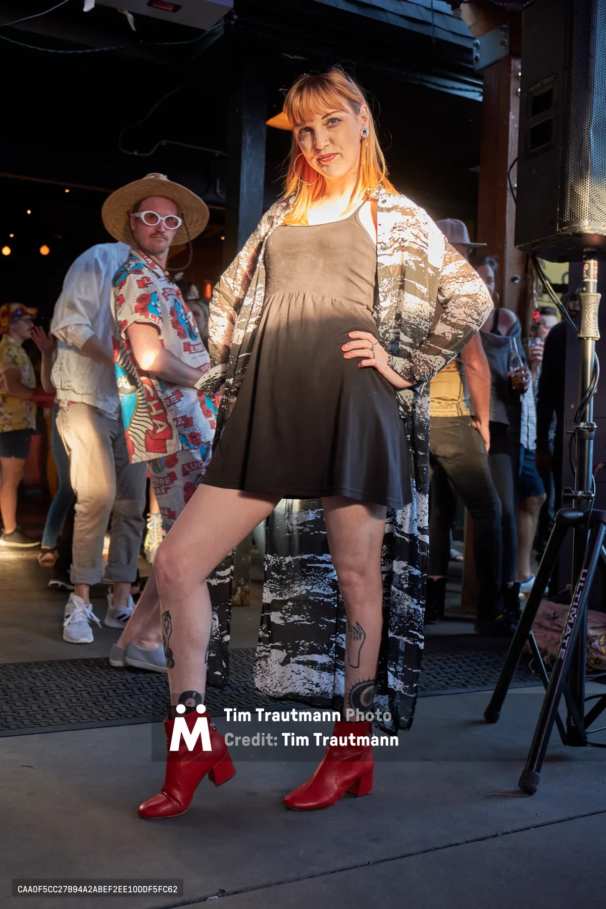 A confident blonde woman poses in the center of White Owl Social Club's industrial interior, wearing a flowing olive jumpsuit layered with a patterned kimono and striking red ankle boots. Behind her, a man in a vintage Hawaiian shirt and straw hat adds to the eclectic summer party atmosphere, while warm string lights illuminate the exposed brick and metal ceiling above. The scene captures the creative energy of Portland's alternative fashion community at this seasonal day party event.