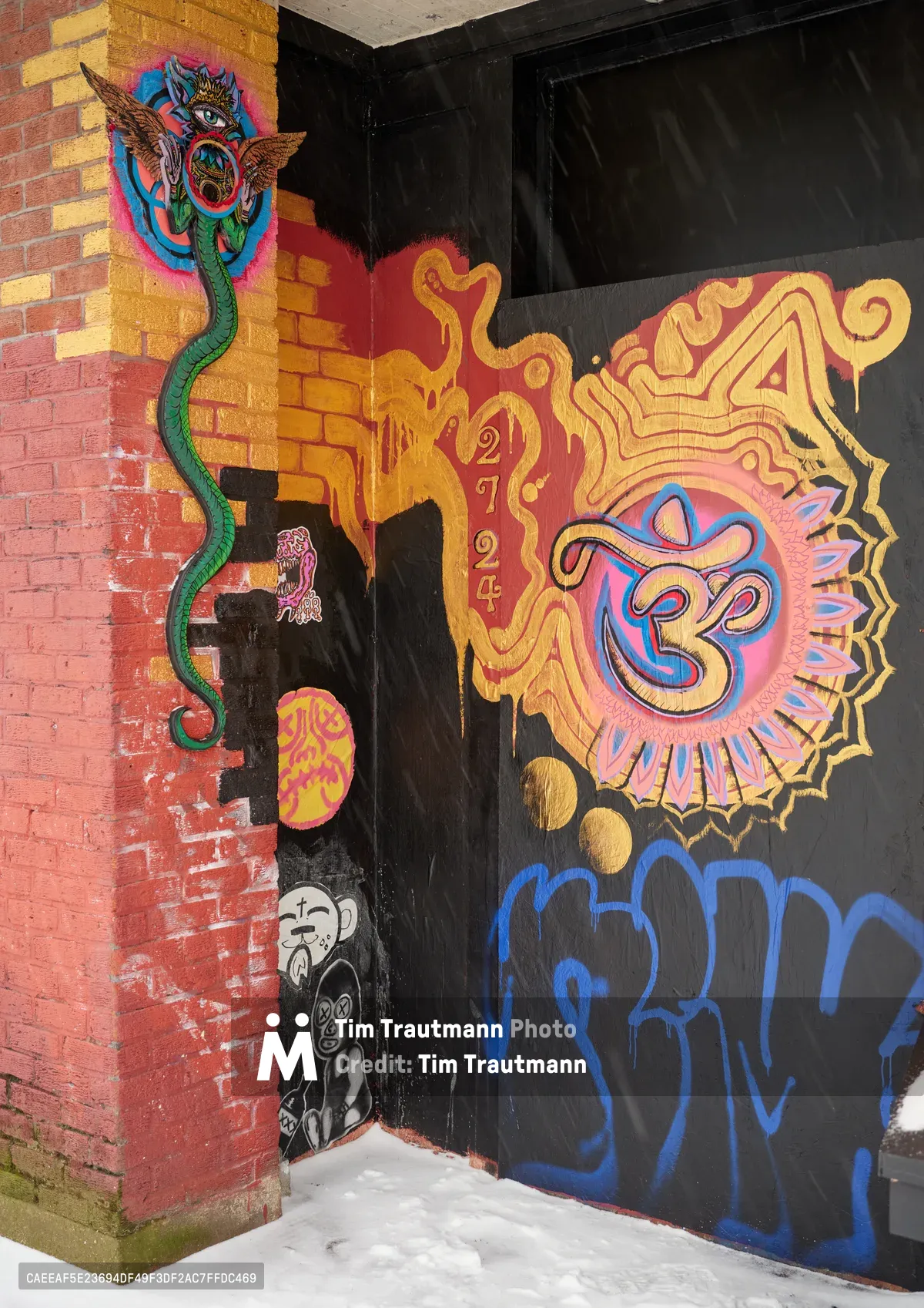 A weathered brick doorway on Northeast Alberta Street becomes a canvas for spiritual street art, where vibrant murals blend Eastern and Western iconography. The composition features a serpentine green dragon coiling around ornate golden lettering, while a radiant Om symbol bursts with pink and blue energy against the dark entrance. Below, a contemplative figure in black and white observes the sacred display, creating a meditation on urban spirituality in Portland's Concordia neighborhood.