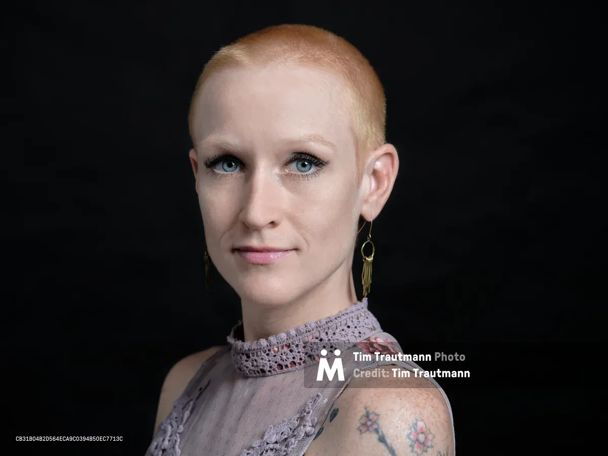 Against the deep charcoal backdrop of a Portland studio, Cayce Ryan commands attention with her striking platinum buzz cut and piercing blue eyes. Her delicate floral top, adorned with intricate lace details at the collar, creates a compelling contrast with her bold personal style and visible tattoo work. The professional headshot captures both vulnerability and strength, with soft studio lighting that emphasizes her direct gaze and the subtle pink tones of her lip color.