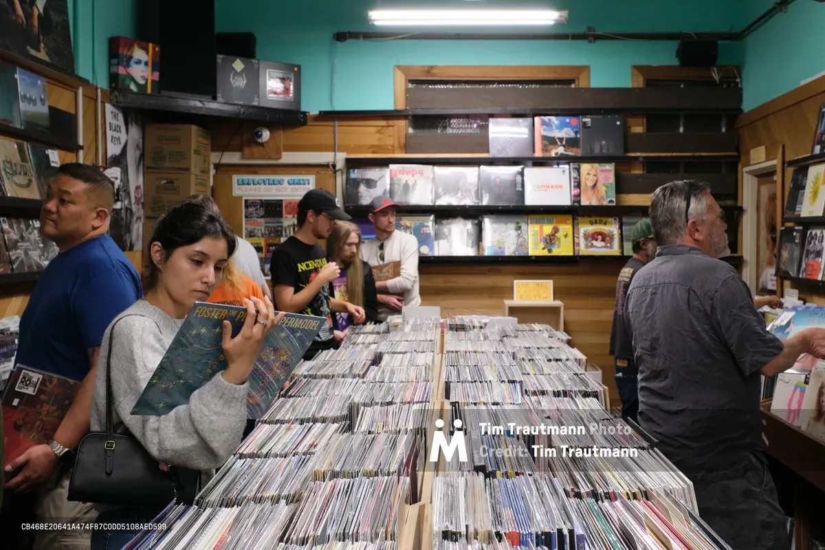 Inside Portland's legendary Music Millennium record store on East Burnside, a young woman in a gray sweater examines vintage vinyl albums while fellow collectors browse through towering bins of records. The turquoise walls and warm fluorescent lighting create an intimate atmosphere as music enthusiasts of all ages dig through the extensive collection. The scene captures the timeless ritual of record hunting in one of the Pacific Northwest's most beloved independent music shops.