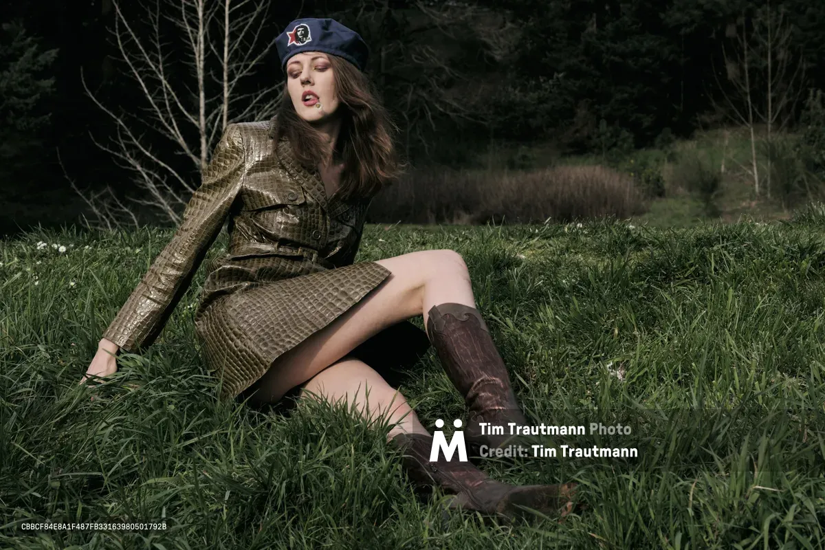 Model Rhianna Atwood reclines gracefully in tall grass wearing a structured olive military-inspired coat and navy beret, captured during golden hour at Mount Tabor Park in Portland, Oregon. The moody lighting creates dramatic shadows across the natural meadow setting, while bare winter trees frame the background. Her contemplative expression and relaxed pose against the earthy terrain evoke a sense of quiet rebellion and seasonal transition.