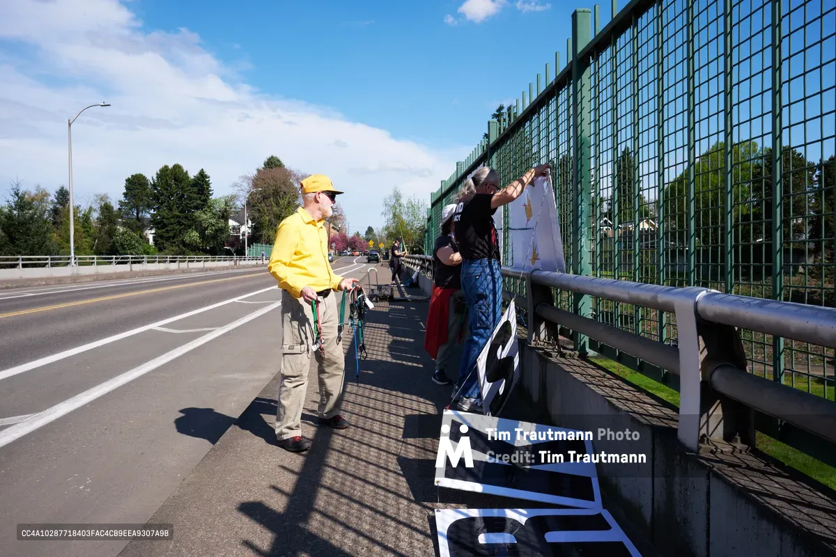 Activists from the Indivisible movement unfurl protest banners along the green mesh fencing of a North Portland overpass, their message advocating for a May 1st General Strike visible to Interstate 5 traffic below. A man in a bright yellow jacket and cap observes as his companion secures white banners bearing bold black text against the afternoon sky. Spring sunlight casts sharp shadows across the concrete walkway, while suburban rooftops and mature trees frame this moment of civic engagement above the busy thoroughfare.