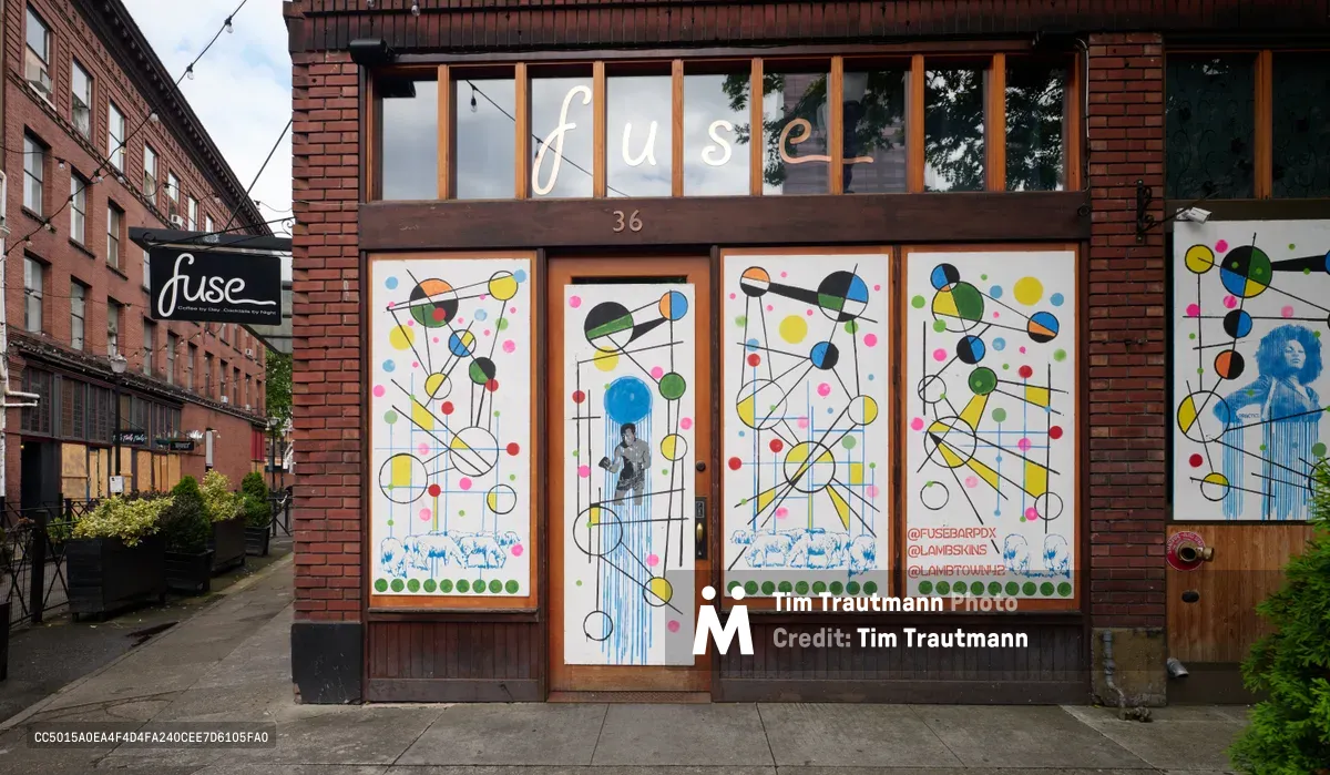 The shuttered facade of Fuse Coffee and Cocktail bar at 36 Southwest Naito Parkway stands transformed by vibrant geometric street art during COVID-19 lockdown. Colorful circles, lines, and dots dance across boarded windows in a kaleidoscope of yellow, blue, green, and pink, while portrait imagery peers through the abstract patterns. The brick building anchors Portland's historic Skidmore/Old Town district, its weathered industrial architecture a stark contrast to the hopeful murals that mask economic uncertainty.
