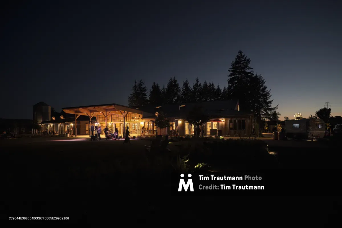 Golden light spills from the windows and covered porches of Abbey Road Farm's rustic buildings as twilight settles over Carlton, Oregon. Silhouettes of guests move across the illuminated terraces while towering evergreen trees frame the pastoral scene against a deep indigo sky. The warm architectural lighting creates an inviting atmosphere that contrasts beautifully with the surrounding darkness of the Willamette Valley countryside.