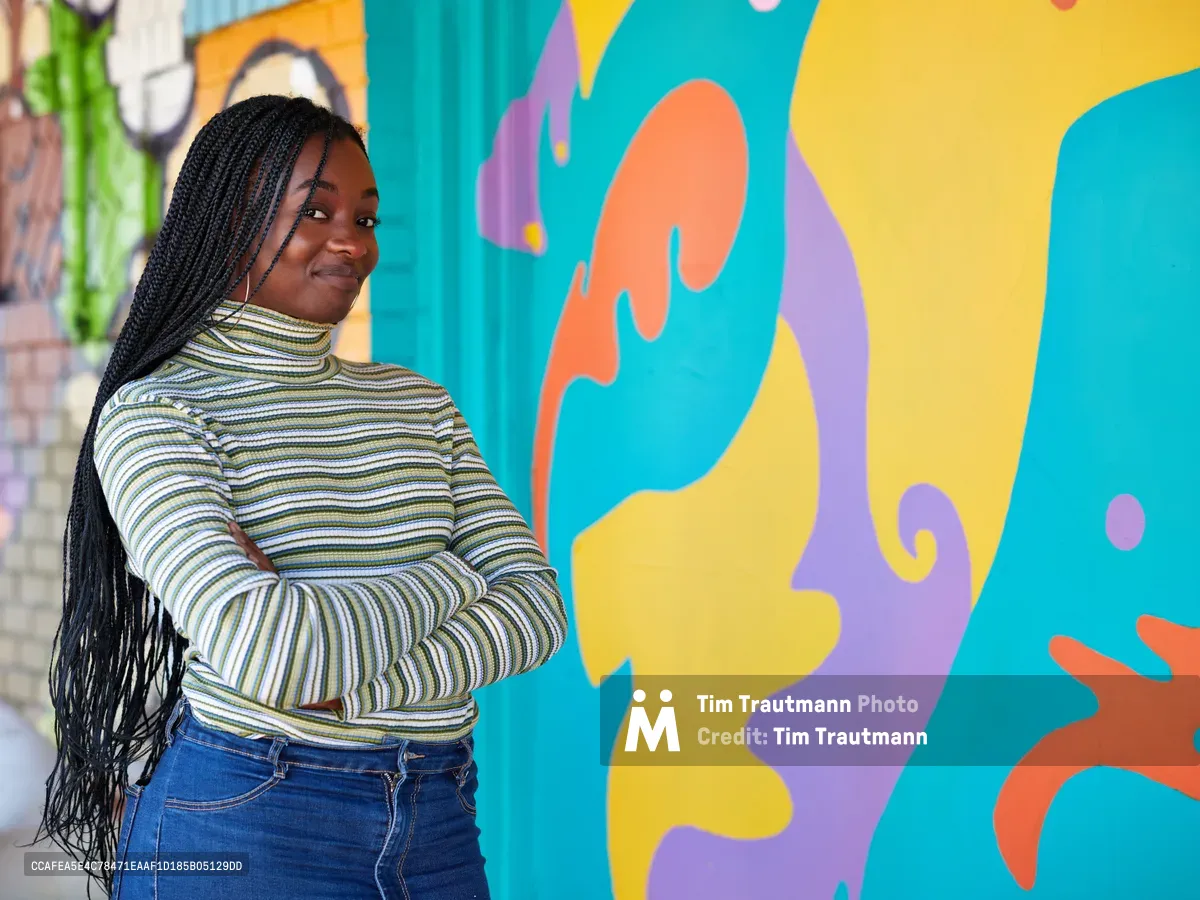 Naomi Likayi stands confidently with crossed arms before her vibrant mural in Portland's Alberta Arts District. The young Black artist wears a striped turtleneck and denim, her long braids cascading as she poses against the flowing abstract forms painted in brilliant turquoise, coral, lavender, and sunshine yellow. The colorful street art transforms the urban wall into a celebration of contemporary African American artistic expression in Northeast Portland.