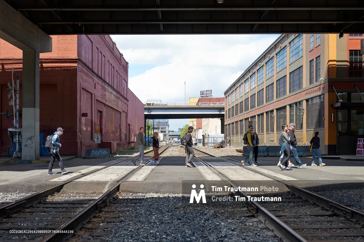 Tour participants navigate active railroad tracks in Portland's Central Eastside industrial district, framed by weathered brick warehouses and converted manufacturing buildings. The scene captures the intersection of urban exploration and industrial heritage, where concrete overpasses span the rails and graffiti-marked walls tell stories of the neighborhood's working-class past. Soft afternoon light illuminates the group as they carefully cross the steel rails, embodying the district's transformation from industrial hub to cultural destination.