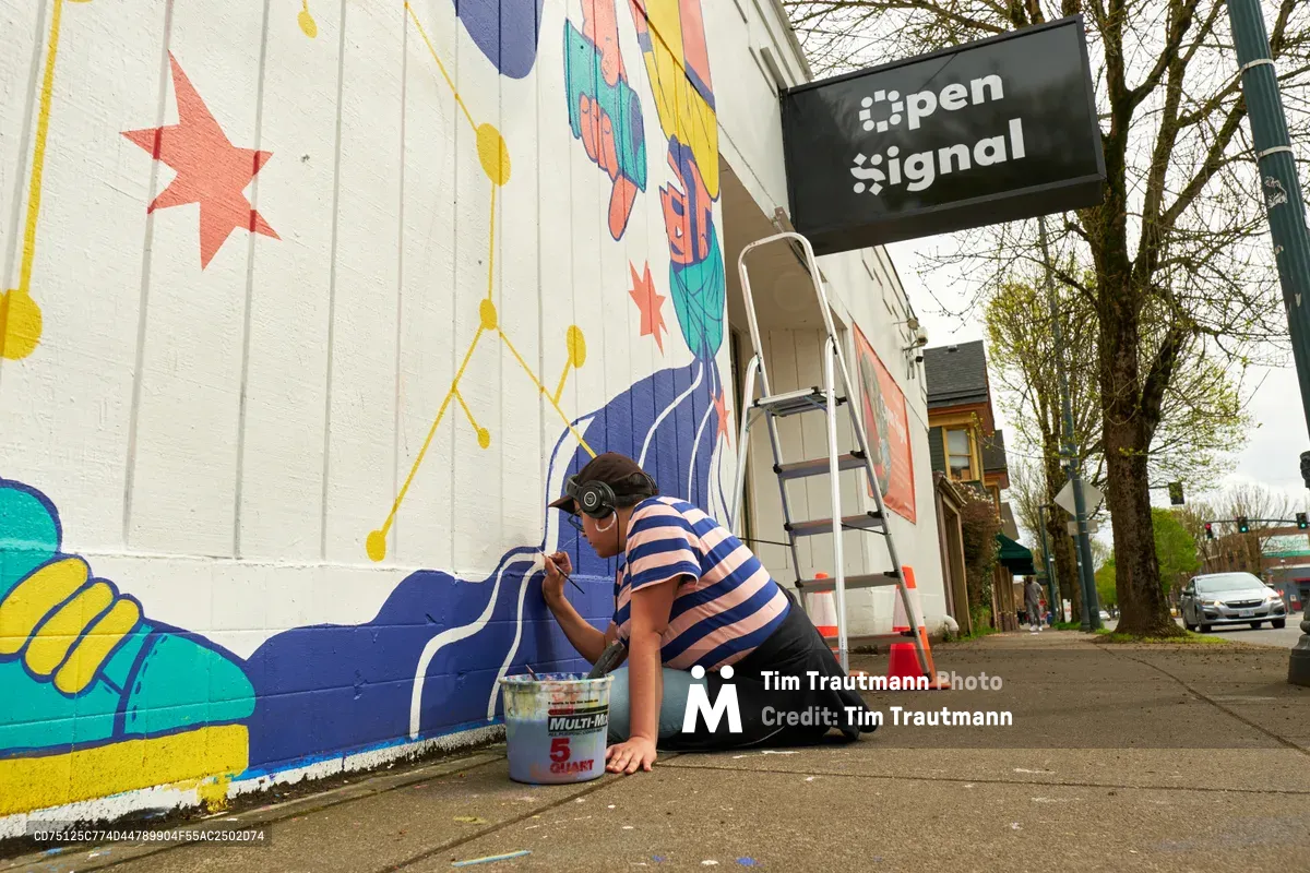 Mexican-American artist Maria Rodriguez, known as Sparkykneecap, kneels on the sidewalk painting vibrant blue details on her commissioned mural "Let's Talk" at Open Signal community media center. Working with focused concentration in a blue and white striped shirt, she applies paint from a bucket while her colorful artwork featuring yellow stars, geometric patterns, and playful figures transforms the white building wall behind her. The spring scene on Northeast Martin Luther King Jr. Boulevard captures the intimate moment of artistic creation within Portland's historic Eliot neighborhood.