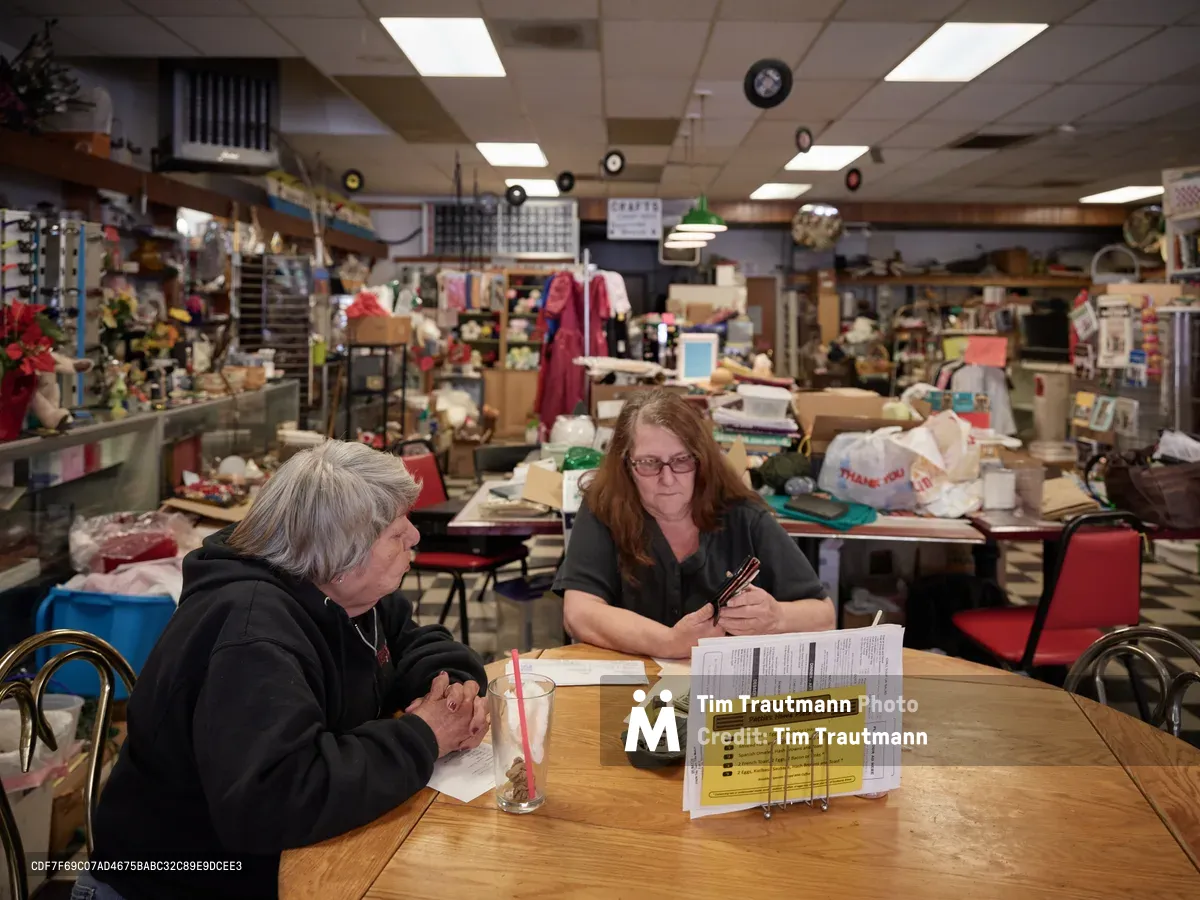 Restaurant owner Pattie Deitz sits across from her best friend Jenny at a worn wooden table in the heart of her bustling North Portland café. The fluorescent-lit interior reveals decades of community gathering, with shelves packed with vintage collectibles, costume jewelry, and local memorabilia creating an intimate backdrop. Scattered paperwork and menus rest between them as they share a quiet moment amid the organized chaos of this beloved Saint Johns neighborhood institution.