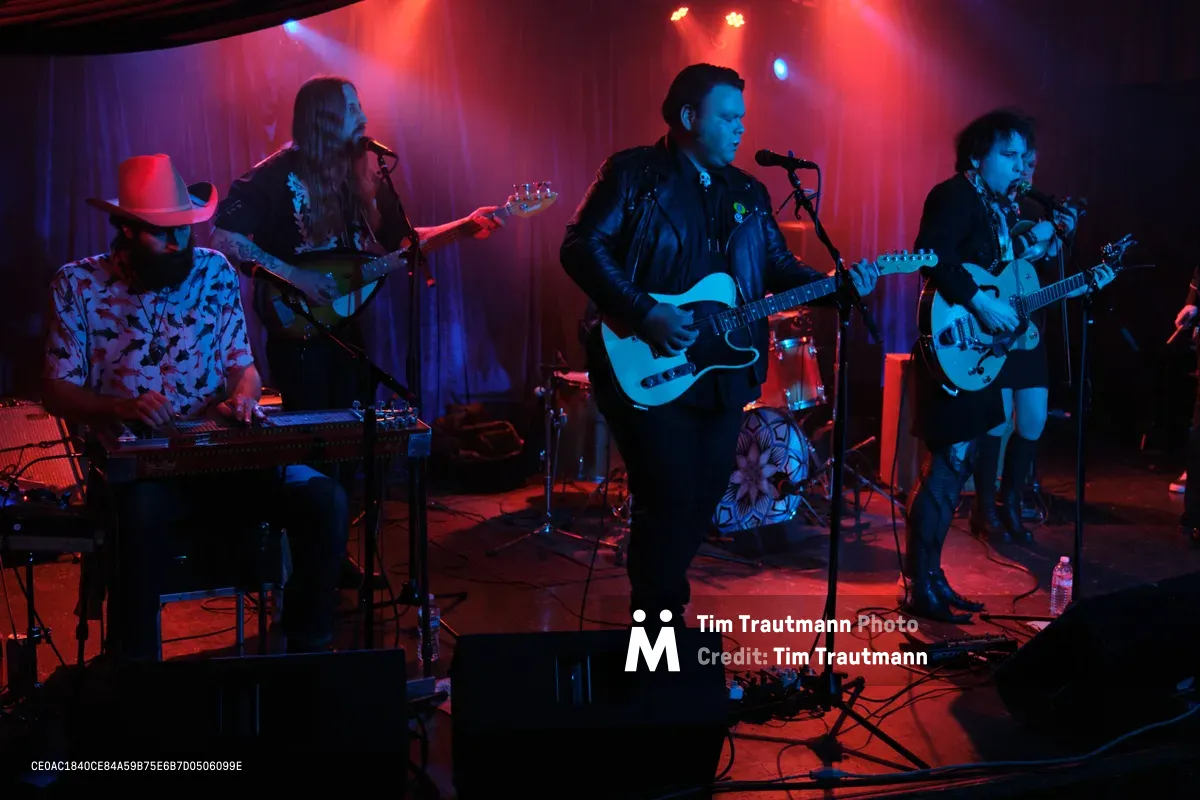The four-piece band Roselit Bone commands the intimate stage at Dante's in Portland's music scene, bathed in dramatic red and blue theatrical lighting. Barry Walker mans the keyboards while wearing a distinctive cowboy hat and patterned shirt, as Tobias Berblinger anchors the rhythm section on bass. Victor Franco delivers vocals while wielding his electric guitar center stage in a black leather jacket, with Charlotte McCaslin adding layers on guitar from stage right. The moody lighting transforms the cramped venue into an atmospheric sonic cathedral, cables snaking across the floor between amplifiers and effects pedals.