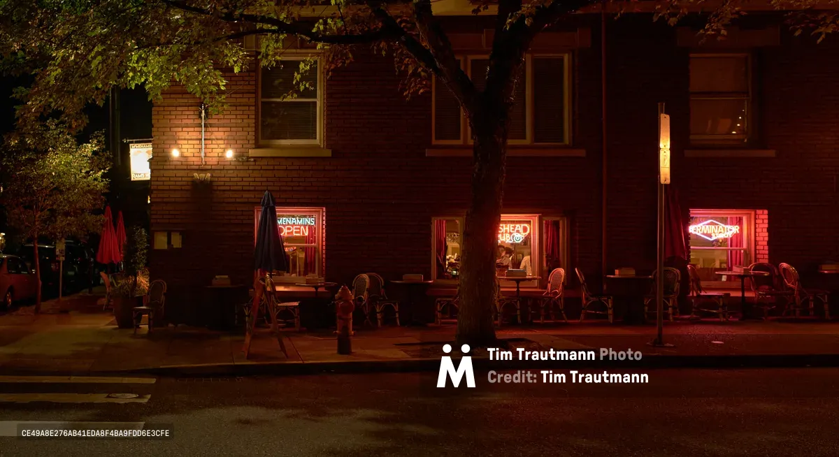 Crimson neon signs illuminate the brick facade of a neighborhood restaurant in Portland's historic Nob Hill district, casting warm red light across empty outdoor seating arrangements. The atmospheric evening scene captures the intimate ambiance of Northwest Glisan Street, where vintage architecture meets contemporary dining culture. Leafy tree branches frame the scene overhead, creating natural shadows that contrast with the artificial glow of the establishment's signage.