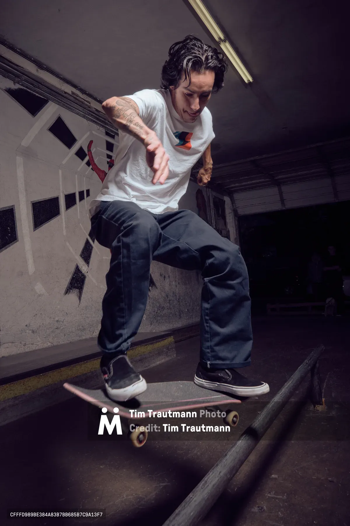A focused skateboarder with tousled dark hair and visible tattoos navigates the gritty confines of Commonwealth Skateboarding's indoor bowl in Portland's Buckman neighborhood. Fluorescent lighting cuts through the shadowed concrete terrain, illuminating his white graphic tee and baggy jeans as he maintains perfect balance on his deck. The raw industrial atmosphere of the Southeast Portland skate shop creates an authentic backdrop for this intimate editorial moment.