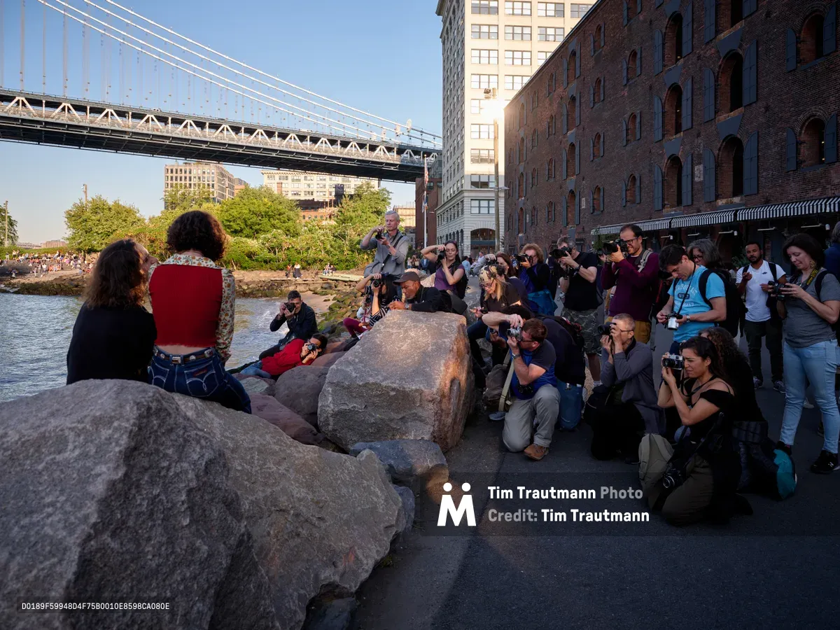 A diverse group of photographers congregates along the rocky shoreline of Brooklyn Bridge Park in DUMBO, their cameras trained on two seated subjects illuminated by golden hour light. The Manhattan Bridge towers majestically overhead while historic warehouse buildings frame the scene. The enthusiastic crowd of amateur and professional photographers creates a dynamic learning environment against the backdrop of New York's iconic waterfront.