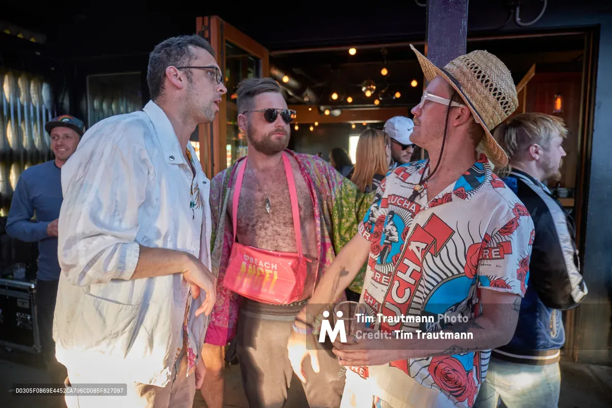 Three men in vibrant summer attire engage in animated conversation during Your Sunday Best's season opener at White Owl Social Club in Portland. The scene captures the eclectic spirit of the daytime party, with one guest sporting a pink mesh tank and bandana, another in a woven hat and patterned shirt, while warm Edison bulb lighting creates an intimate atmosphere against the venue's industrial backdrop. The candid moment embodies the relaxed social energy of Portland's alternative summer scene.
