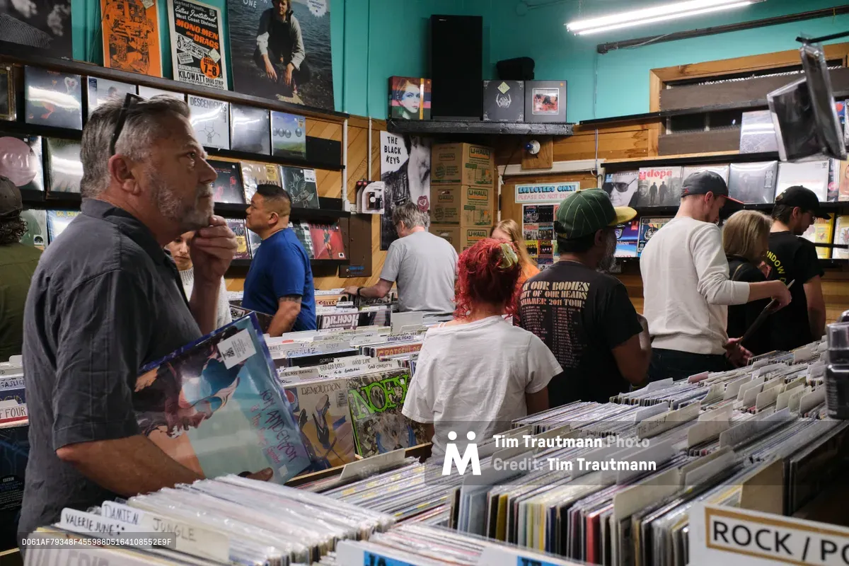 Inside the iconic Music Millennium record store on East Burnside Street in Portland, Oregon, vinyl enthusiasts of various ages dig through extensive collections organized in wooden bins. Fluorescent lighting illuminates the mint-green walls adorned with vintage concert posters and album covers, while a bearded man in the foreground examines a record sleeve. The cramped quarters buzz with the quiet intensity of serious collectors hunting for rare finds among thousands of alphabetically sorted albums.