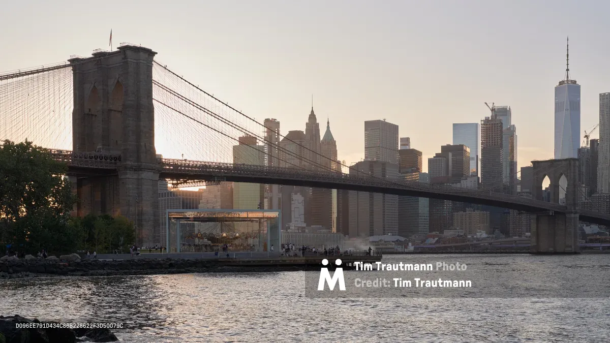 The iconic Brooklyn Bridge spans majestically across the East River as viewed from Brooklyn Bridge Park in DUMBO, with Manhattan's towering skyline creating a dramatic backdrop. Golden hour light bathes the Gothic Revival stone towers and suspension cables in warm amber tones, while the Financial District's skyscrapers, including One World Trade Center, pierce the hazy evening sky. Gentle ripples catch the fading light on the river's surface as visitors gather along the waterfront promenade to witness this quintessential New York vista.