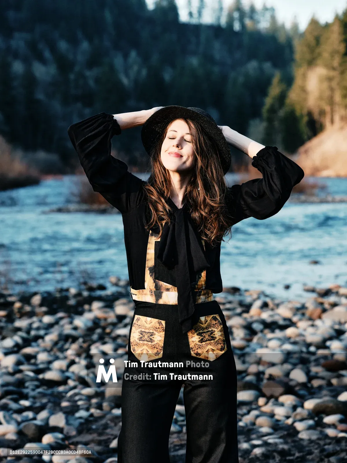 A young woman with flowing chestnut hair stands in peaceful contemplation along Oregon's Floodplain Trail, her arms gracefully raised to adjust her black wide-brimmed hat. She wears a distinctive black ensemble adorned with golden leaf-pattern accents that echo the autumn landscape behind her. The rocky riverbed of what appears to be the Sandy River stretches beneath her feet, while soft natural light illuminates her serene expression against the backdrop of dense Pacific Northwest forest.