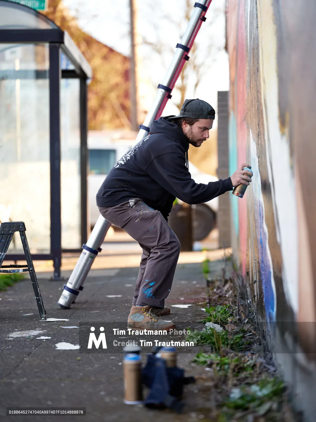 Travis Fields, known as campographic, works intently on a vibrant mural depicting poet Amanda Gorman at the intersection of Northeast 15th Avenue and Killingsworth Street in Portland's Alberta district. The bearded artist in dark hoodie and backwards cap stretches toward the wall with spray can in hand, his extension ladder propped nearby. Scattered paint cans dot the weathered pavement while soft afternoon light illuminates this urban canvas transformation in one of Portland's culturally rich neighborhoods.