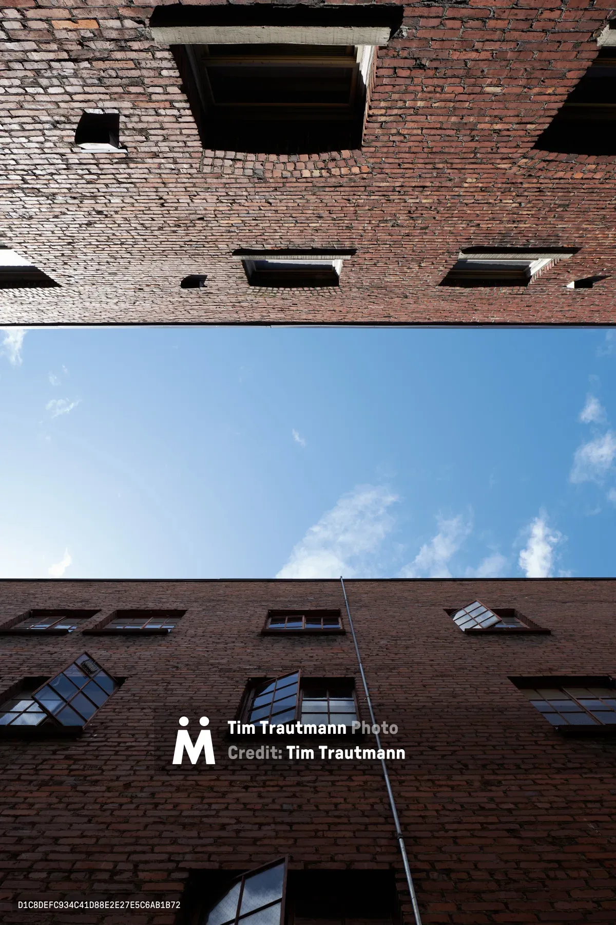 A dramatic worm's-eye view captures the towering brick walls of a historic warehouse courtyard in Portland's Central Eastside Industrial District. The weathered red brick facades frame a narrow slice of azure sky dotted with white clouds, while industrial windows reflect the afternoon light. The geometric composition emphasizes the imposing height and industrial heritage of this converted warehouse space on Southeast Yamhill Street.