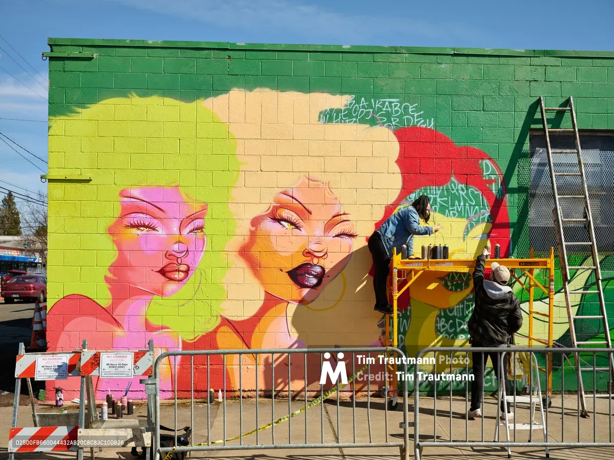 Multidisciplinary artist Vilatose works from scaffolding to paint a vibrant pop art mural titled 'Jardín Cubana' featuring three female figures on the green brick wall of a building in Portland's Alberta Arts District. The artist applies paint with careful precision while safety barriers protect the work zone below, capturing the creative energy of Northeast Alberta Street's thriving street art scene under clear blue skies.
