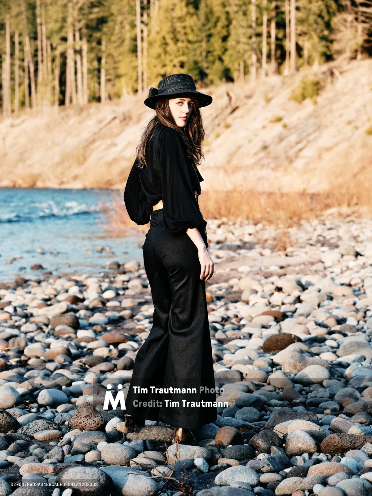 A woman in flowing black attire and wide-brimmed hat stands contemplatively among the weathered river stones of Oregon's Floodplain Trail. Golden hour light bathes the pebbled riverbed and distant forested hillsides in warm amber tones, creating an atmosphere of quiet introspection. Her silhouette against the natural landscape evokes a sense of solitary wandering and connection with the Pacific Northwest wilderness.
