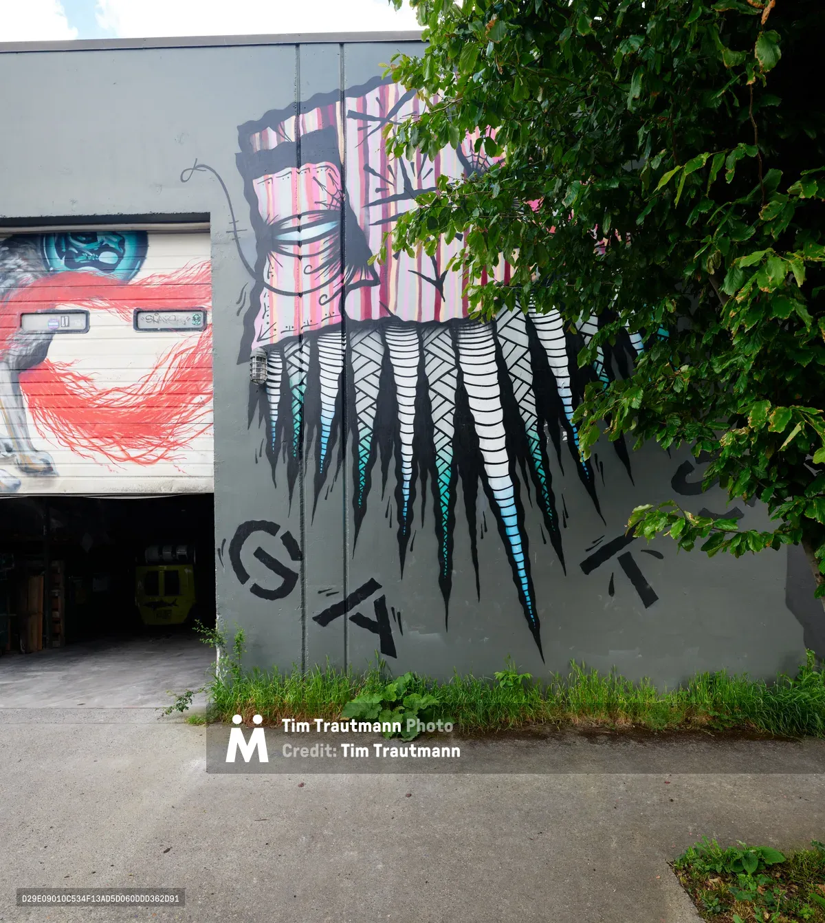 A vibrant street mural transforms a concrete wall in Portland's Kerns neighborhood, where cascading feathers in turquoise and coral tones flow beneath overhanging green foliage. The artwork depicts a striking eye peering through red brushstrokes, while geometric patterns and stylized text create layers of urban expression against the industrial gray facade. Summer light filters through the leafy canopy above, creating dappled shadows that dance across the painted surface.