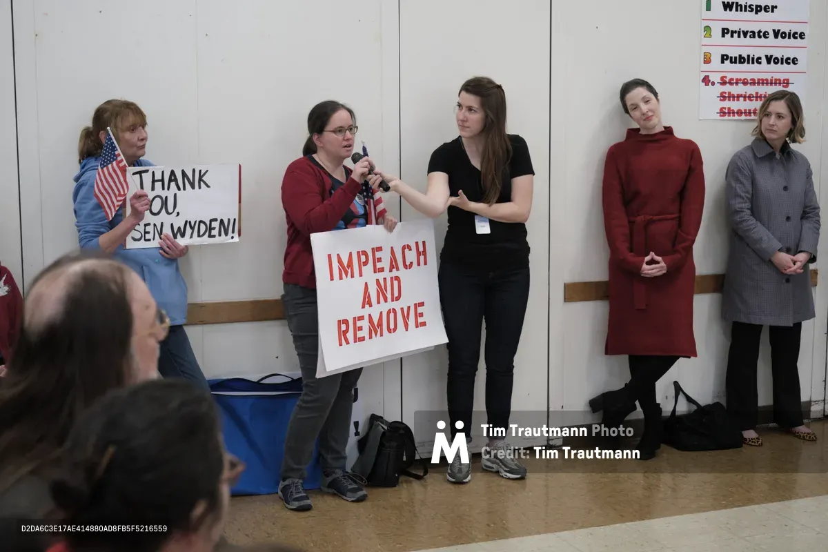 Constituents gather in the fluorescent-lit multipurpose room of Robert Gray Middle School in Portland's Hillsdale neighborhood, wielding handmade protest signs during Senator Ron Wyden's town hall meeting. A woman in glasses and burgundy cardigan commands the microphone while another holds an "IMPEACH AND REMOVE" placard, as three other attendees stand silently against the institutional white walls. The scene captures the raw energy of grassroots political engagement, with everyday citizens transforming a school cafeteria into a forum for democratic discourse.