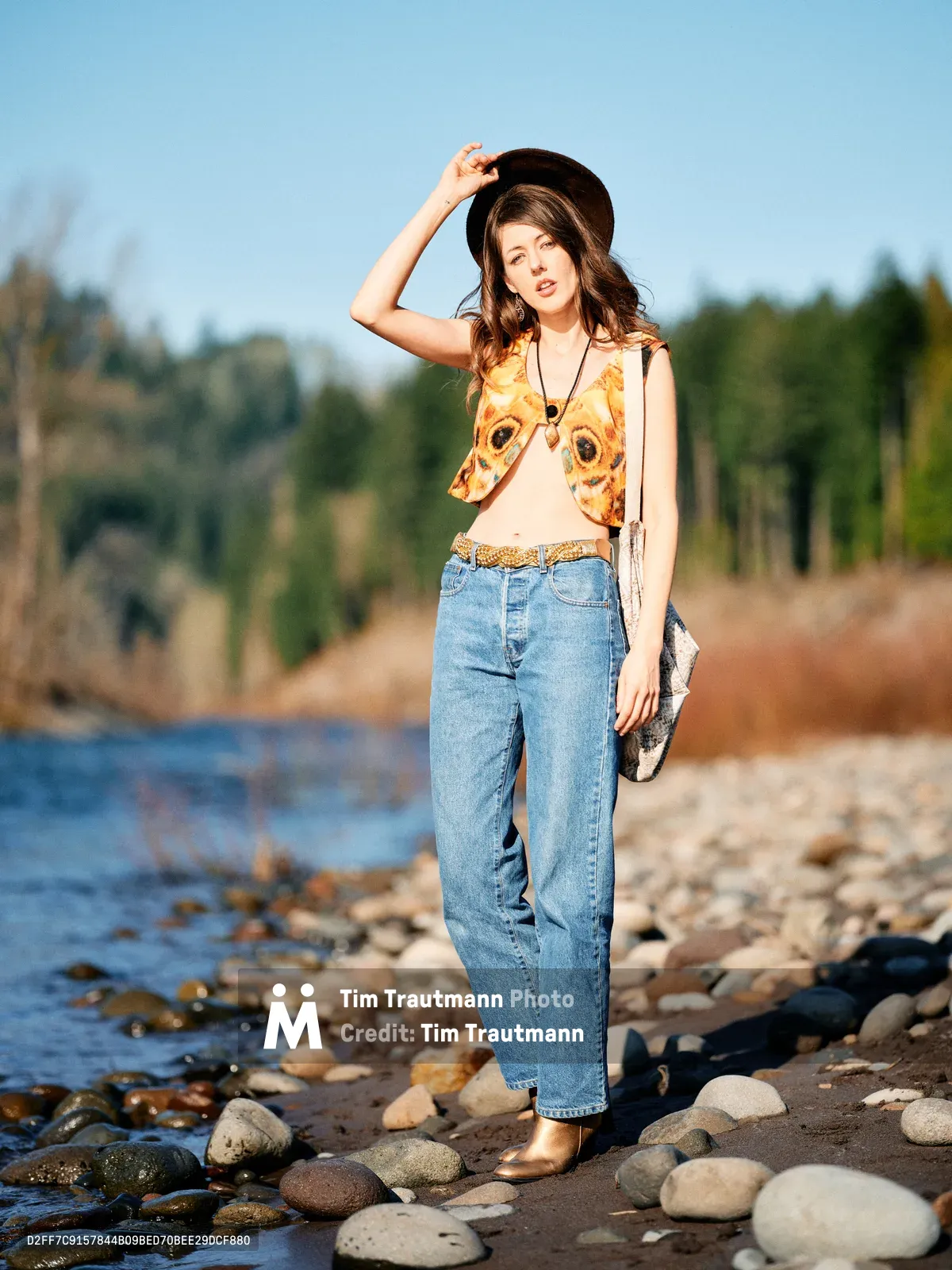 A young woman in vintage-inspired attire stands confidently among smooth river stones along the Floodplain Trail in Multnomah County, Oregon. She wears a distinctive animal-print vest over a cream tank top, high-waisted denim, and a dark wide-brimmed hat, her hand gracefully positioned at the hat's brim as she gazes toward the camera. The golden hour light bathes the scene in warm tones, while the gentle waters and forested hills of the Pacific Northwest create a serene backdrop. Her bohemian styling and the natural setting evoke a sense of wanderlust and connection to the Oregon wilderness.