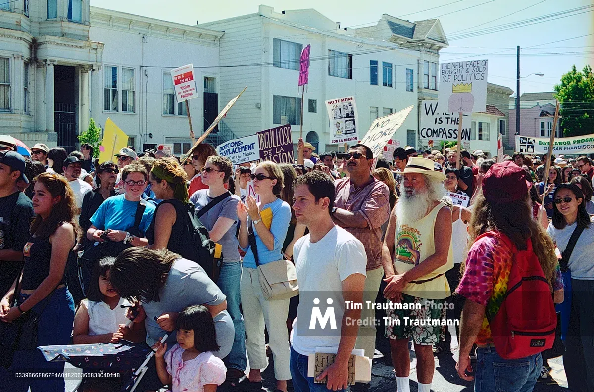 A diverse crowd of hundreds fills the sunny streets of San Francisco's Mission District during an anti-displacement demonstration organized by the Mission Anti-Displacement Coalition. Protesters of all ages carry handmade signs and banners opposing gentrification, with Victorian architecture and white apartment buildings creating a distinctly San Francisco backdrop. The bright midday sun illuminates the passionate faces of residents fighting to preserve their neighborhood's affordability and cultural character against the pressures of tech industry expansion.