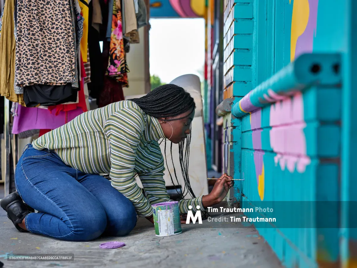 Naomi Likayi kneels on the sidewalk outside a vibrant clothing boutique, carefully signing her completed mural on turquoise-painted brick walls. Her striped long-sleeve shirt and concentrated expression capture the intimate moment of artistic completion in Portland's Alberta Arts District. Colorful fabrics and garments hang in the shop window behind her, creating a rich tapestry of community commerce and creative expression.
