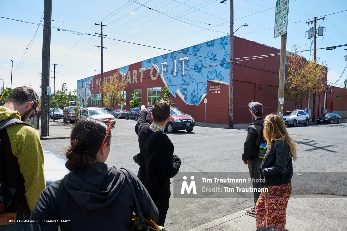 A diverse group of participants gathers on Southeast Taylor Street in Portland's Central Eastside district, their attention focused on a vibrant mural adorning a red brick industrial building. The afternoon sun illuminates the scene as tour guide Tomás Valladares leads an interpretive discussion about the neighborhood's street art culture. Power lines create geometric patterns overhead while parked cars and urban infrastructure frame this moment of cultural discovery and community engagement.