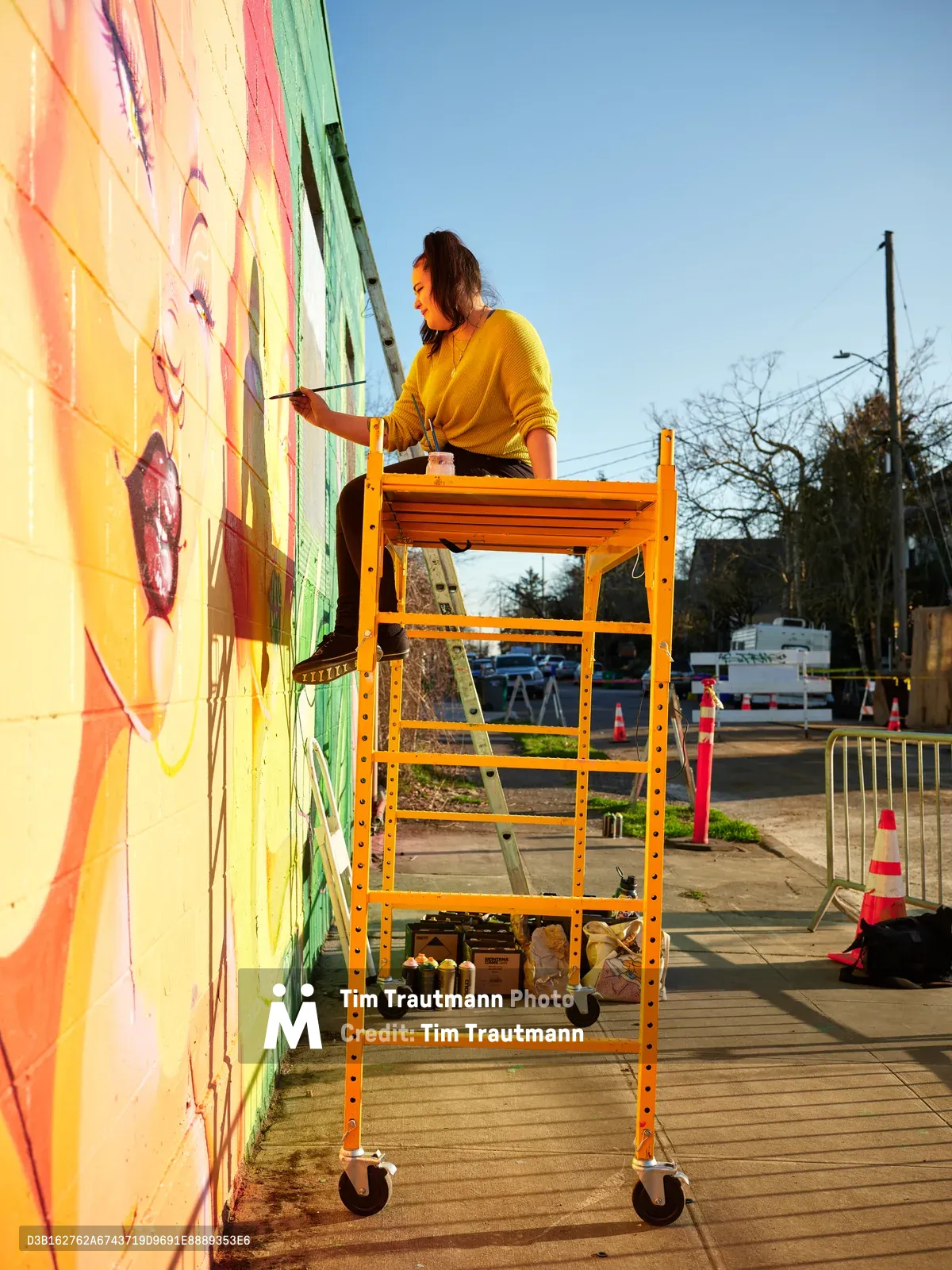 Multidisciplinary artist Villatose works from an orange rolling scaffold against the brilliantly colored mural 'Jardín Cubana' on Northeast Alberta Street in Portland's Alberta Arts District. Golden hour light bathes the scene as the artist, wearing a yellow sweater, applies brushstrokes to the vivid composition featuring three female figures in green, pink, and yellow. Traffic cones and street barriers frame the urban workspace, while bare winter trees and power lines create geometric patterns against the clear blue sky.