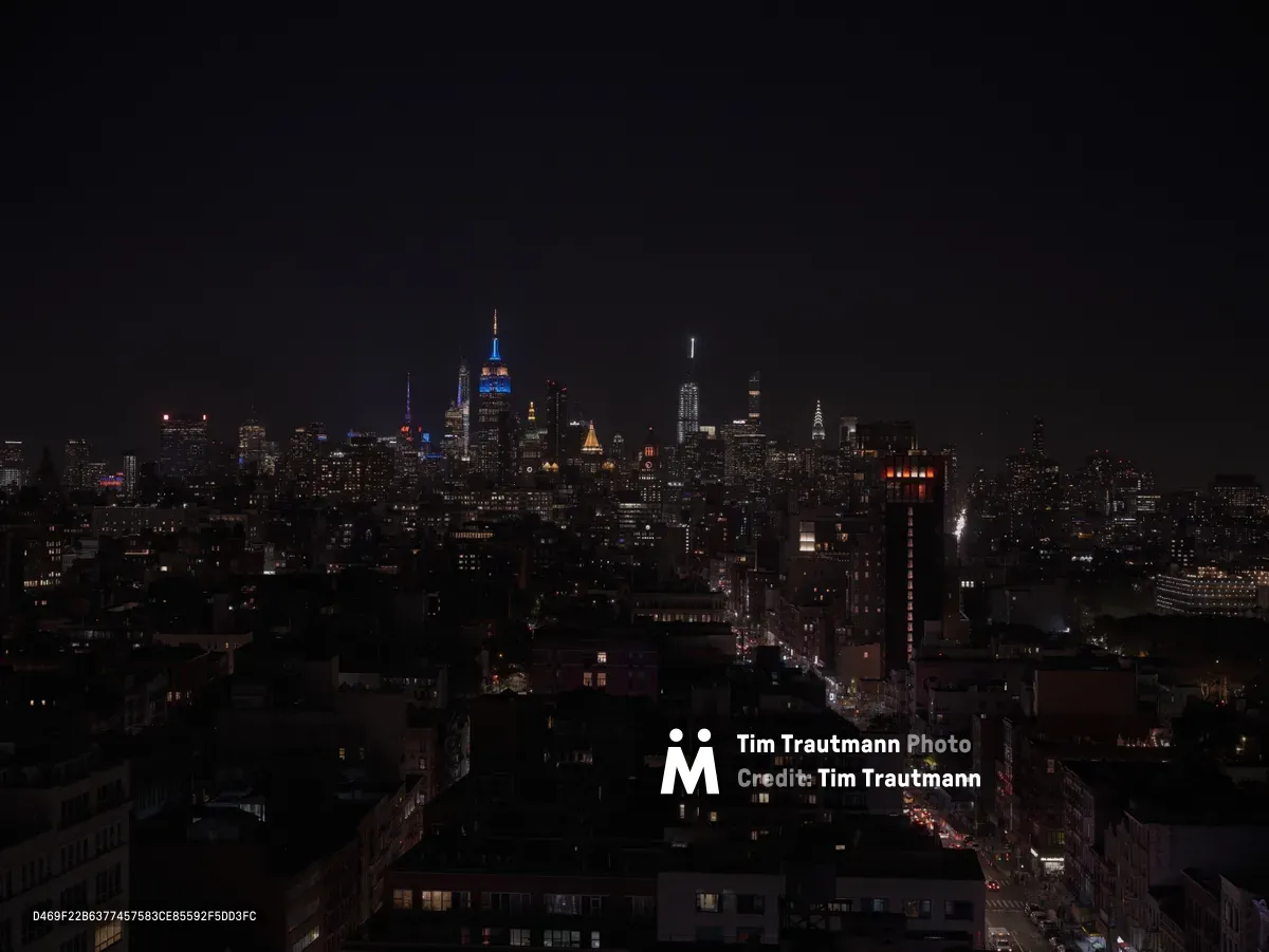 The iconic Manhattan skyline glows against an inky black sky, photographed from the elevated perspective of Canal Arcade in Chinatown. The Empire State Building dominates the distant midtown cluster, its illuminated spire piercing the darkness while countless office windows create a constellation of warm light across the urban landscape. Street-level traffic streams through the foreground neighborhoods, their headlights tracing arterial pathways that connect the intimate scale of Lower Manhattan's residential blocks to the towering commercial heart of the city. The composition captures the dramatic contrast between the intimate human scale of Chinatown's low-rise buildings and the soaring ambition of Manhattan's architectural monuments.
