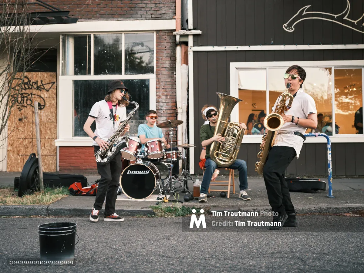 A spirited brass quartet brings jazz to the sidewalk during Last Thursday's street fair on Alberta Street in Portland's Concordia neighborhood. Two saxophonists flank the ensemble while a tuba player and drummer anchor the rhythm section, their instruments gleaming in the late afternoon light. The urban backdrop of weathered brick buildings and storefront windows frames this impromptu concert, capturing the authentic spirit of Portland's monthly art walk where music spills onto the pavement and community gathers around creativity.