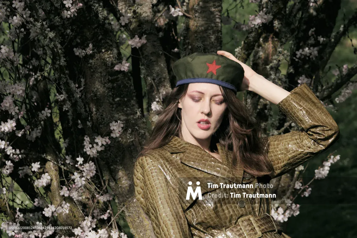 Model Rhianna Atwood poses contemplatively among blooming cherry blossoms at Mount Tabor in Portland, Oregon, wearing a golden crocodile-textured leather coat and olive military cap adorned with a red star. The soft spring light filters through the delicate pink sakura petals, creating an evocative juxtaposition between militant iconography and nature's gentle renewal. Her hand rests thoughtfully against the cap's brim as she gazes downward, surrounded by the ephemeral beauty of hanami season.