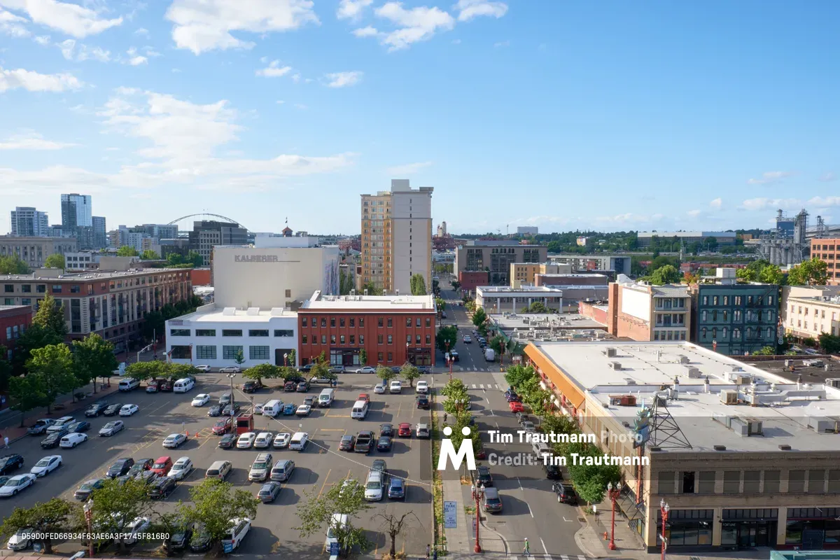 An elevated afternoon view captures the bustling intersection of Northwest Portland's Pearl District, where historic brick buildings meet contemporary architecture under a canvas of scattered cumulus clouds. The scene reveals a well-organized street grid with tree-lined avenues, surface parking lots filled with vehicles, and the distinctive mix of red brick facades and modern glass structures that define this revitalized neighborhood. Golden hour light bathes the urban landscape, highlighting the textural contrast between the weathered industrial buildings and sleek residential towers rising in the distance.