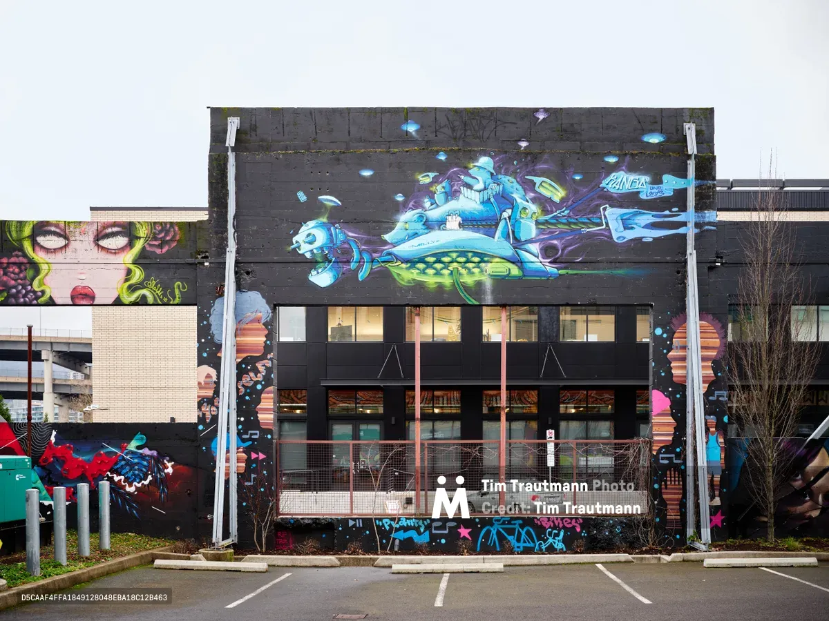 A striking street art mural dominates the black facade of Clay Creative in Portland's Hosford-Abernethy neighborhood, featuring a vibrant underwater scene with flowing sea creatures in electric blues, purples, and greens. The fantastical aquatic imagery, commissioned by the Portland Street Art Alliance, transforms the former Taylor Electric building into a canvas of liquid motion and marine mythology. Warm interior lighting glows through the building's windows beneath the dramatic artwork, while overcast Pacific Northwest skies create a muted backdrop that makes the mural's luminous colors pop with ethereal intensity.